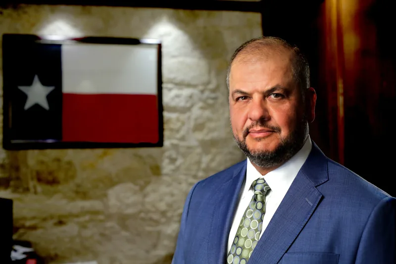 Ernest Acevedo III in a blue suit with a Texas flag behind him